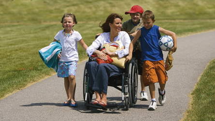 Children pushing a woman in a wheelchair with some groceries