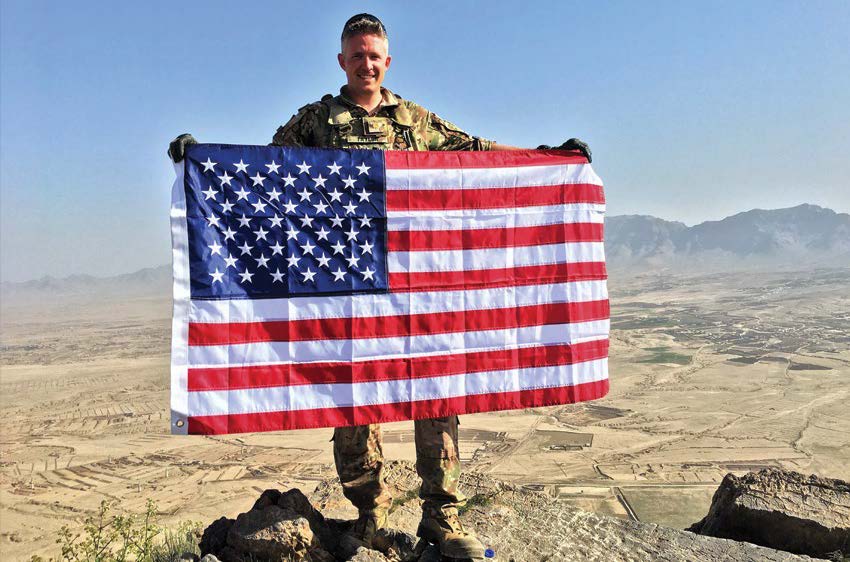 : Major Brent Taylor unfurling an American flag on a mountaintop in Afghanistan during the summer of 2018. Courtesy of Jennie A. Taylor
