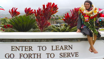 A female student wearing several leis sitting on the welcome sign to BYU Hawaii