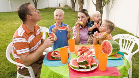 A family sitting at a table in the back yard eating watermelon